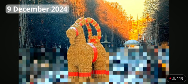 The Gävle Goat on the morning of 9th of December 2024.

Picture shows a large straw effigy of a goat with red trimmings surrounded by a fence. Behind the goat a tree lined esplanade and the Gävle theatre are visible. Parts of the image are pixelated to obscure people passing by.

Picture from YouTube Live Feed. 