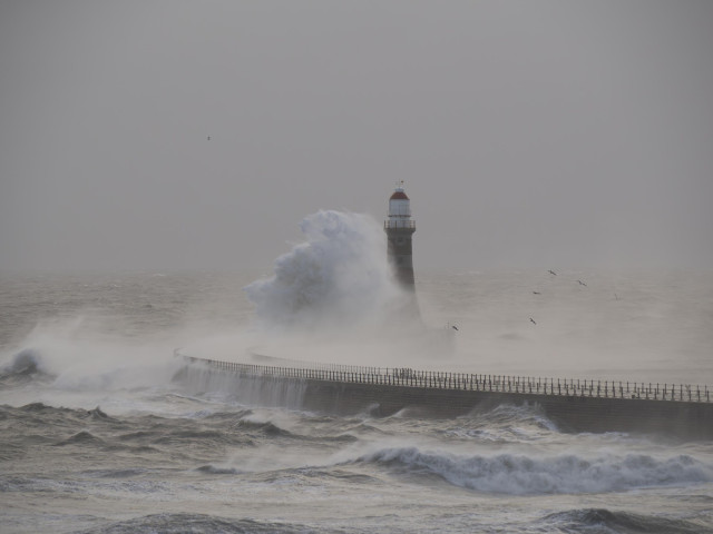 A large wave hits the pier and creates a wall of water that rises and engulfs the Roker Lighthouse