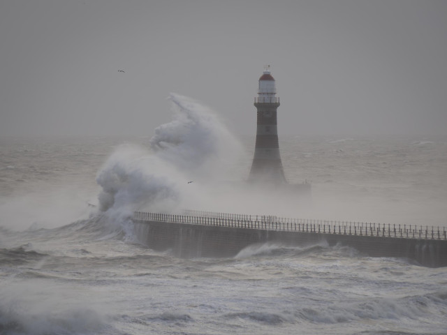 A large wave hits the pier and creates a wall of water that rises and engulfs the Roker Lighthouse