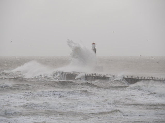 A large wave hits the pier and creates a wall of water that rises and engulfs the Roker Lighthouse