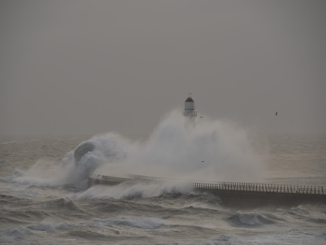 A large wave hits the pier and creates a wall of water that rises and engulfs the Roker Lighthouse