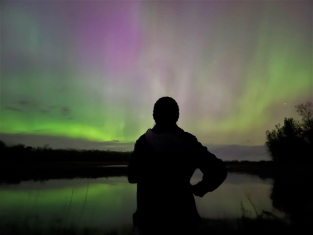 Aurora photo of a woman standing in silhouette with green and purple aurora above reflected in the water of a pond in front of her. May 10, 2024. 10:49 pm. Flynn Township, Michigan. May 10, 2024. 10:49 pm. Flynn Township, Michigan.
