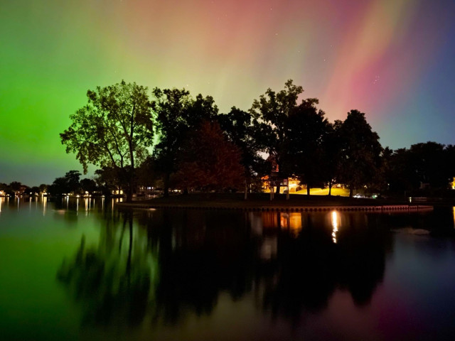 Aurora photo of a rainbow of bright covers over an island in a suburban lake. The trees of the island and the colors are reflected in the lake in the foreground. Oct. 10, 2024, 10:06 pm. Wolverine Lake, MI.
