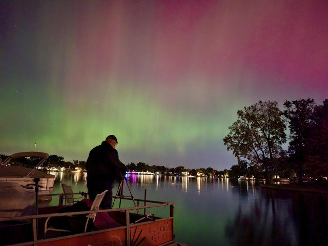 Aurora photo of green and red aurora over a suburban lake. In the left foreground a man on an orange pontoon boat is adjusting a camera on a tripod. Oct. 10, 2024, 10:18 pm. Wolverine Lake, MI.
