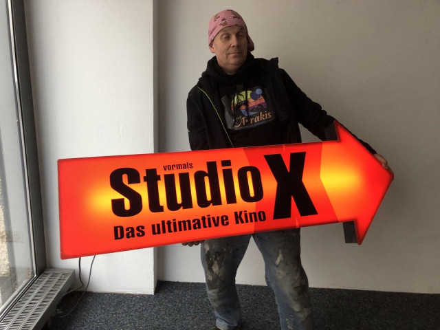 I'm holding the arrow of an orange and red neon sign in my arms. It's about 1.5 metres long and has the words 'StudoX - the ultimate cinema' written on it