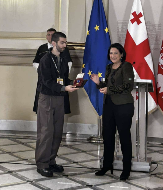 President of Georgia awarding a medal to journalist in a civic ceremony in front of flags of Georgia and the E.U.