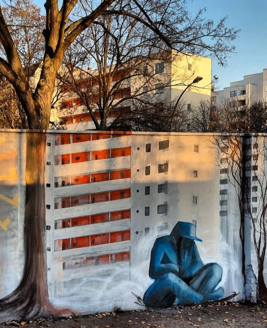 Streetartwall. A socially critical mural was sprayed/painted on a white street wall and incorporates the surroundings behind it. A blue male figure with a cap can be seen on the wall, slumped on the ground. He seems to be freezing, but a face is not recognizable. Next to him we see the façade of a high-rise building with red balconies and the trunks of two trees, painted to match the actual surroundings behind the wall. Behind the wall are the tree and a white skyscraper with red balconies. Some of the trees are not in the picture to better show off the building behind it. A touching mural about the sometimes invisible poverty in a big city.