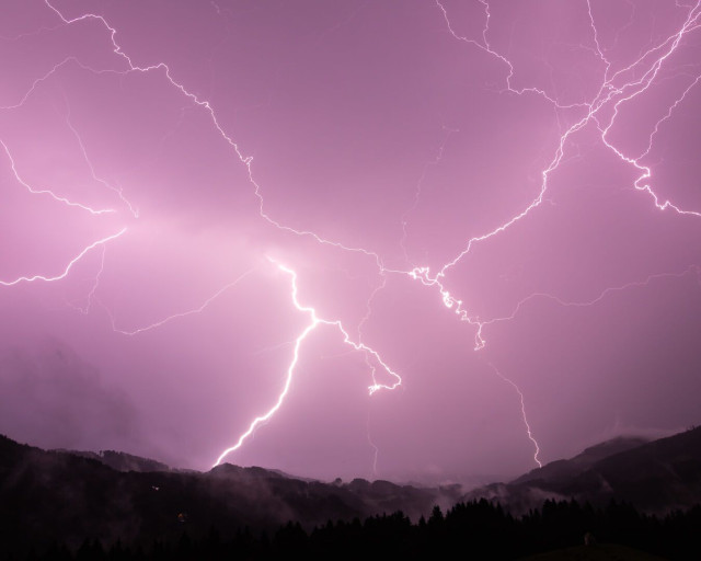 In violet-rot gefärbte verschiedenen große Blitze vor einem bewölkten Nachthimmel. Am unteren Ende des Bildes ist die Silhouette einer Hügelligen Waldlandschaft zu erkennen.