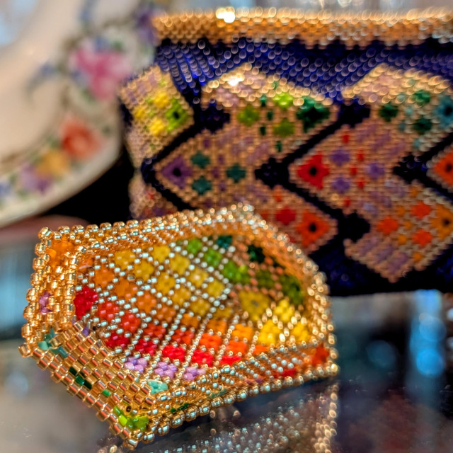 Photograph of the interior of a hexagonal box lid made out of 11/0 beads. The color design is a diagonal rainbow gradient computer of diamond shapes outlined in gold. The box itself, which has golden rainbow fish against a deep navy blue, is visible in the background. CC BY-SA Kate Zimmerman from a pattern by Julia Pretl.