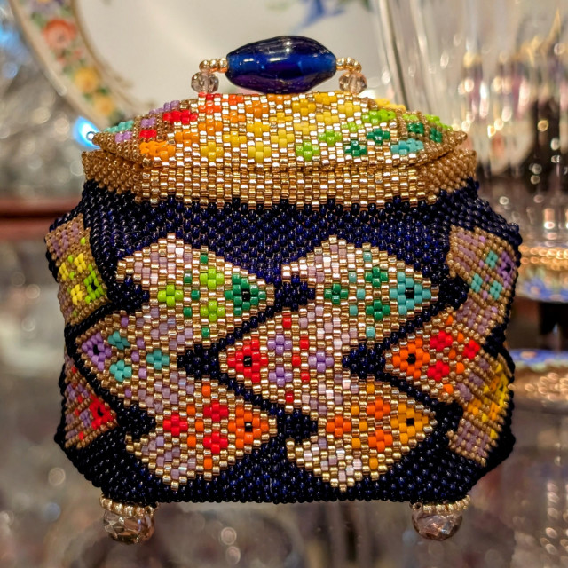 Photograph of a hexagonal box made out of 11/0 beads. This is my favorite of the beaded boxes I made using Julia Pretl's "Little Bead Boxes" patterns. The background uses deep navy blue translucent Czech beads. The fish and the lid are composed of Delica beads, outlined in gold, with the interiors using different colors of the rainbow. Mapping out how I wanted the rainbow colors to progress around the box took some time. CC BY-SA Kate Zimmerman.