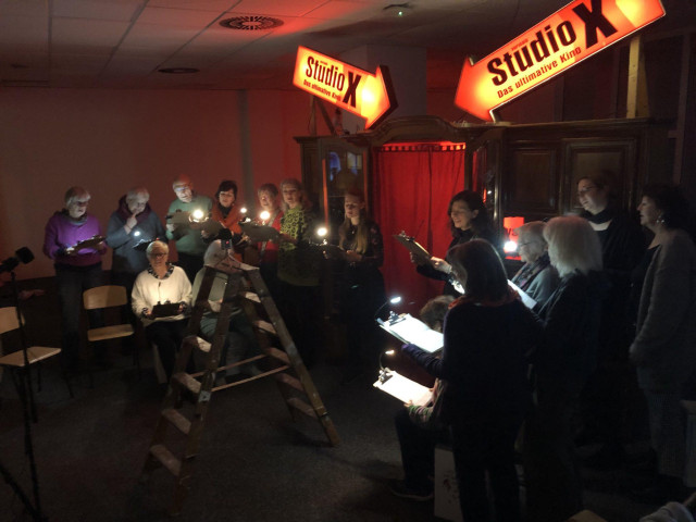 about twenty persons are standing in front of a livingroom closet. They have closet with lights in their hands and are rehearsing a speaker performance. The cupboard has been converted into a kind of confessional. At the top of the cupboard is a neon sign for a porn cinema