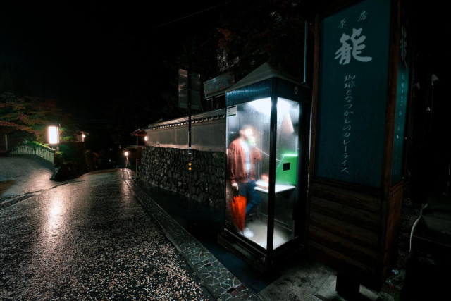 A photo of a street on a rainy night, and a phone booth. A person can be seen inside the phone booth, and they have a red umbrella by their leg. The glass is completely fogged up, so it's hard to see any details inside the phone booth.