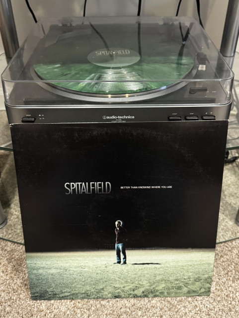 A turntable with a marbled green vinyl record playing, overlaid with the album cover for "Better Than Knowing Where You Are" by Spitalfield. The album cover features a figure standing alone in a barren landscape.