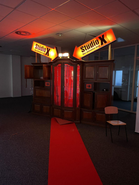 A living room cupboard in the Gelsenkirchen Baroque style stands in a showroom. It has been converted into a walk-in object. The centre section, which juts out like a bay window, has become a cabin. Inside it glows red. What is hidden inside is concealed by a red curtain. At the top of the cabinet are two arrows from a cinema neon sign pointing to the centre section. A red carpet leads to the wardrobe 