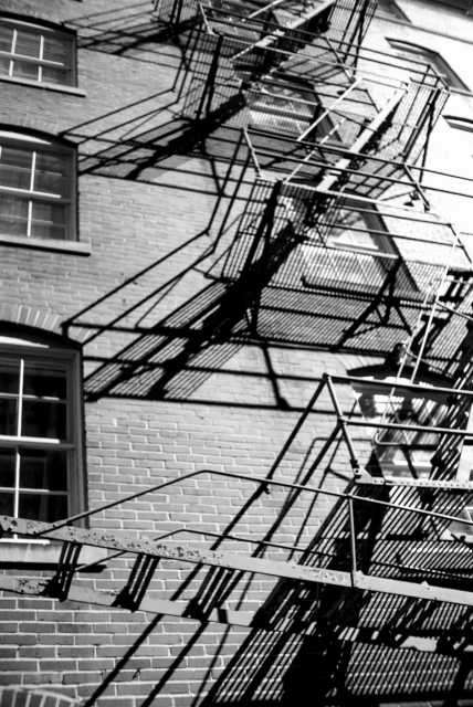 A black and white photo looking up the side of a building at a series of fire escapes whose shapes are creating an interesting pattern of lines