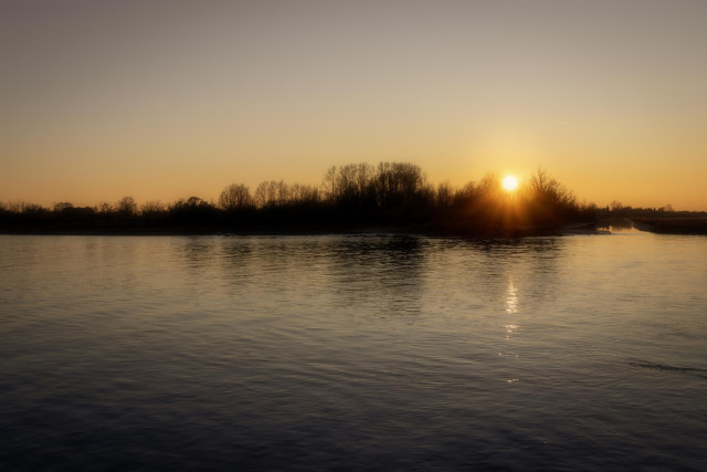 Fotografía donde un poco más de la mitad inferior se ve agua con reflejos de luz y árboles. En la mitad superior se ven las siluetas de árboles y el sol, que se está poniendo, asomando entre ellas.