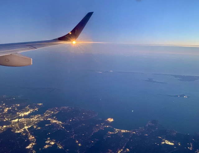 A plane wing is visible against a twilight sky, with the sun setting in the background. Below, a view of land with city lights and a body of water is seen, depicting a scenic aerial landscape.