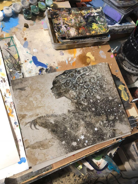 An early photo of the figure in progress with the face defined in silhouette. Taken at an angle, the artist's illustration table occupies most of the photo. The space is a bit of a mess. This is how the sausage is made!
