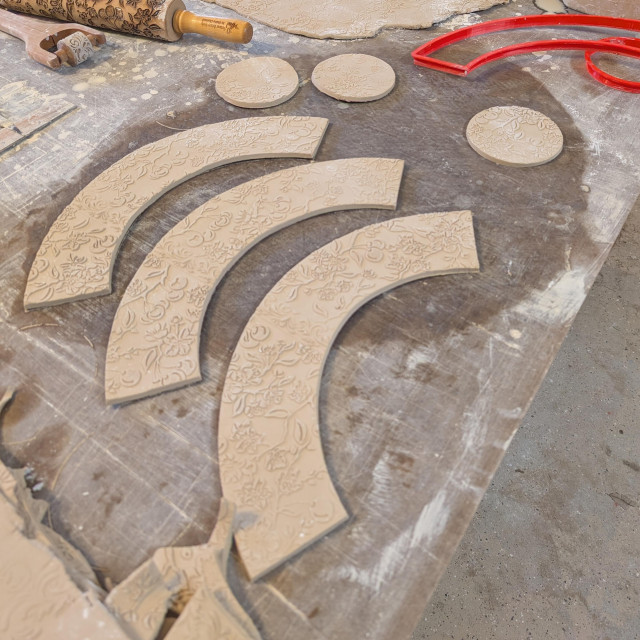 A slab of clay rolled out, textured and cut into almost rainbow-like shapes that can be formed into bowls. The texture is cherries, cherry blossoms, and small branches with leaves. Also on the table is a pile of clay leavings, the rolling pin used to make the texture, and the cutter used to actually cut the shape. 