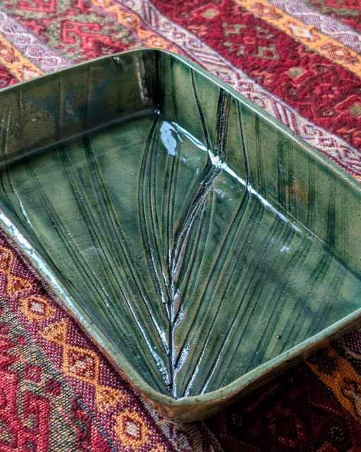 Photograph of a deep green rectangular dish, texture coming from the speckled buff clay and the imprinted palm frond. The color is from a black copper oxide wash with clear glaze on top. CC BY-SA Kate Zimmerman.