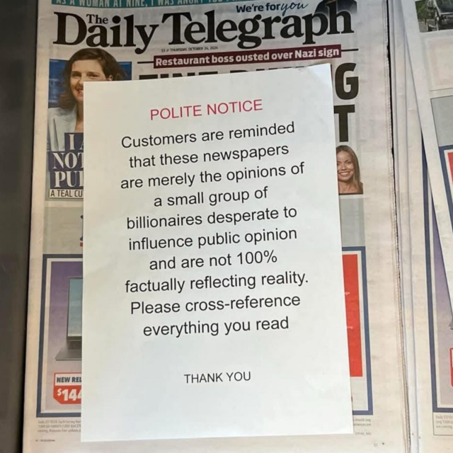 A Daily Telegraph newspaper on a stand. Over the top of it an A4 sized piece of paper has been placed. The text on the paper reads:

POLITE NOTICE

Customers are reminded that these newspapers are merely the opinions of a small group of billionaires desperate to influence public opinion and are not 100% factually reflecting reality.

Please cross-reference everything you read!

THANK YOU