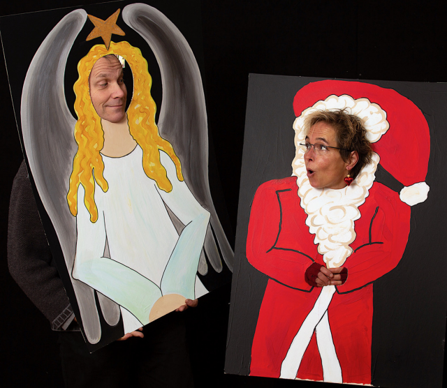 Two people stand in front of a black background and look at each other. Each person is looking through an opening in a cardboard box. An angel is painted on one cardboard box and a Father Christmas on the other. The place where the figures' heads should be is an opening through which you can stick your head. So it looks as if the angel and Father Christmas are looking at each other