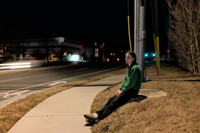 Night. A woman in a green shirt, black pants, and sandals sits on a short hill by the sidewalk of a wide road. A streetlamp illuminates her vicinity, and a nearby intersection's signals surround her head. An oncoming car's headlights blur from the long exposure.