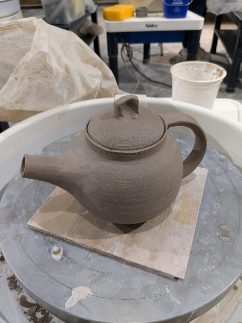 A small teapot sitting on a pottery wheel. It has a stubby spout and a pulled handle. The lid fits nicely, at least.