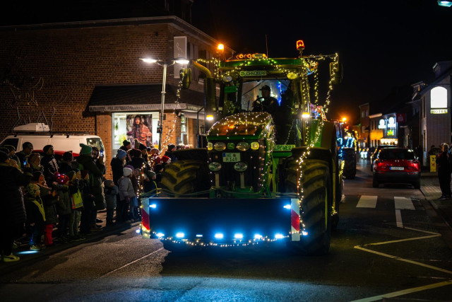 Ein weihnachtlich beleuchteter Traktor fährt durch ein Dorf, am Wegesrand sind viele Menschen zu sehen. 