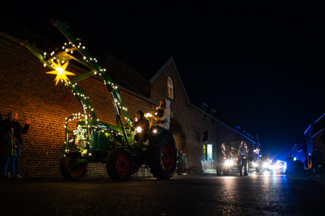 Ein beleuchteter Traktor fährt durch ein Dorf, dahinter sind die Frontlichter von anderen Fahrzeugen zu sehen. 