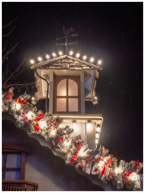 A decorative rooftop with a small gazebo features warm lights and festive garlands adorned with red ribbons. A weather vane sits atop, and gentle rain adds a cozy ambiance to the night scene.