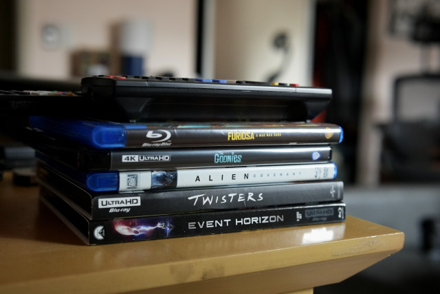 A remote on top of a stack of Blu-rays and 4K UHD Blu-rays.  Furiosa: A Mad Max Saga (BD), Goonies (4K), Alien Covenant (BD), Twisters (4K) and Event Horizon (4K).