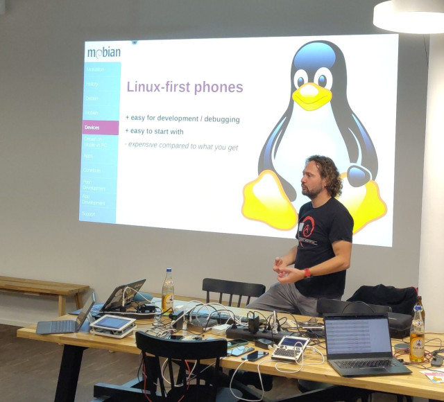 Slide about linux-first phones with a big on the right side. The presenter is wearing a black debian shirt. On the desk in front of the speaker many mobile devices, and two club mate bottles.