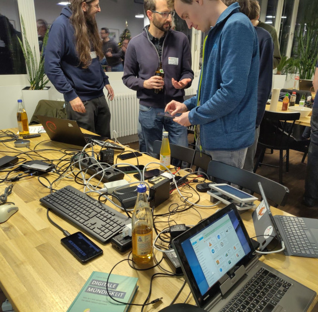 A table with many mobile linux devices: smartphones and tablets, a laptop and a industrial terminal. Besides that some USB chargers and a lot of cables, three bottles, two of them are club mate and book called 'Digitale Muendigkeit'. Next to the desk 6 people, some of play around with smartphones, one is holding a beer. Plants in the background.