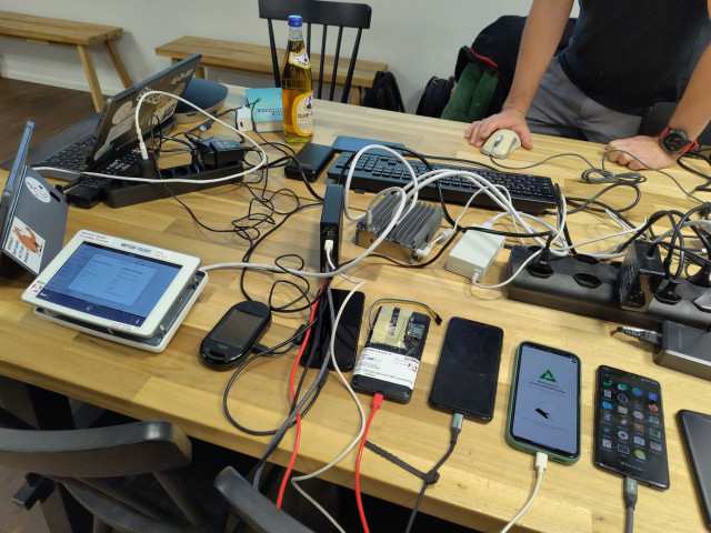 A table with at least 7 phones, running mobile Linux, two convertibles a METTLER TOLEDO industrial terminal and a lot of cables. A keyboard and mouse. Club Mate.