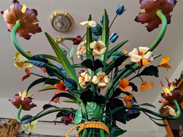 Photograph of a painted tole chandelier seen from the bottom, with branches and flower sprays arching  upward. The leaves and stems are painted in varying shades of green, the flowers range from yellow to orange to pink to purple to blue. CC BY-SA Kate Zimmerman.