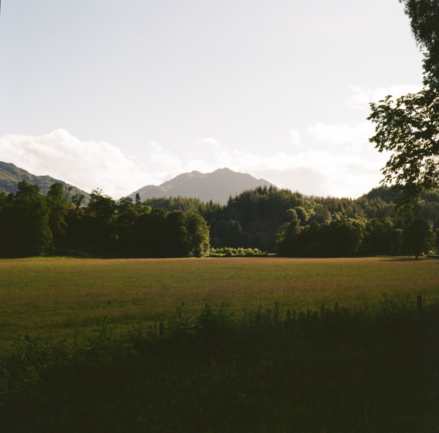 A colour photograph of a late evening scene on summer solstice looking over fields, trees and a mountain 