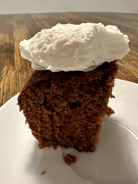 A slice of gingerbread sitting on a white saucer. It has a dollop of whipped cream on top like a hat. 