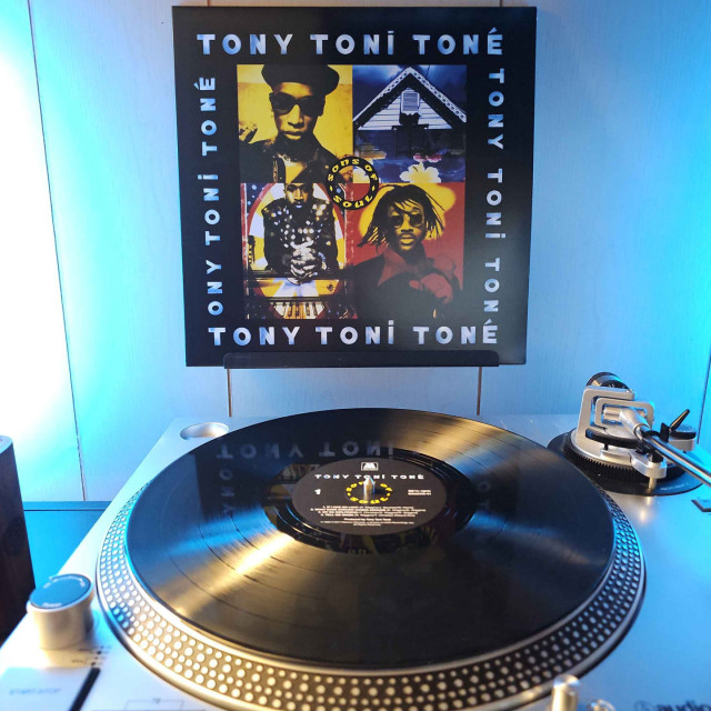 A vinyl record sits on a turntable. Behind the turntable, a vinyl album outer sleeve is displayed. The front cover shows 4 quadrants. 3 feature a different group member, and the fourth on the upper right shows the upper floor of a house from the outside . "Tony! Toni! Toné" is on each side of the cover. 