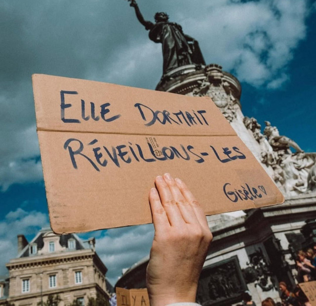 hand raising a poster at protest against sexual violence, blue marker on cardboard, saying 
'elle dormait
réveillons - les
Gisèle ♡'