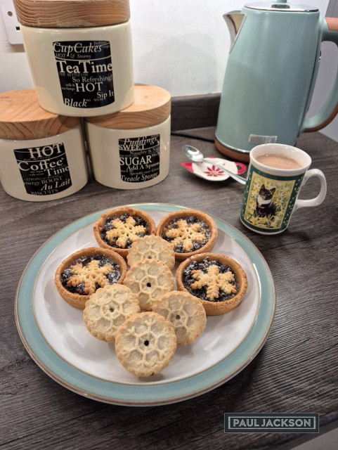 A mug featuring a charming cat design sits on the kitchen counter. Beside it are five mini mince pies on a plate with light golden pastry with tops shaped like snowflakes and a dusting of sugar. On the same plate are four larger mince pies with shortcrust pastry cases that are open to show the generously filled mincemeat infused with brandy and cider.