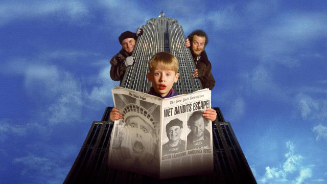 Kevin McAlister with a newspaper open. Front page says the Wet Bandits Escape. Behind him is the Empire State Building. Harry and Marc are behind Kevin. 