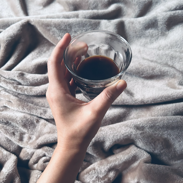 A square photo in colour. The photo shows a soft, light-tan blanket over which Maitreya's left hand, also light-tan in hue, though a different shade than the blanket. Her hand is holding out a small, clear-glass, bevelled glass with black coffee, & the inside side of the glass has slight condensation from the steam coming from the hot coffee. Gentle light is coming in from the left of the frame, from where the window is, & the photo is lit only by natural, indirect light from a partly-cloudy morning outside. The moment is cosy & content, with a sense of comfort & relaxation.