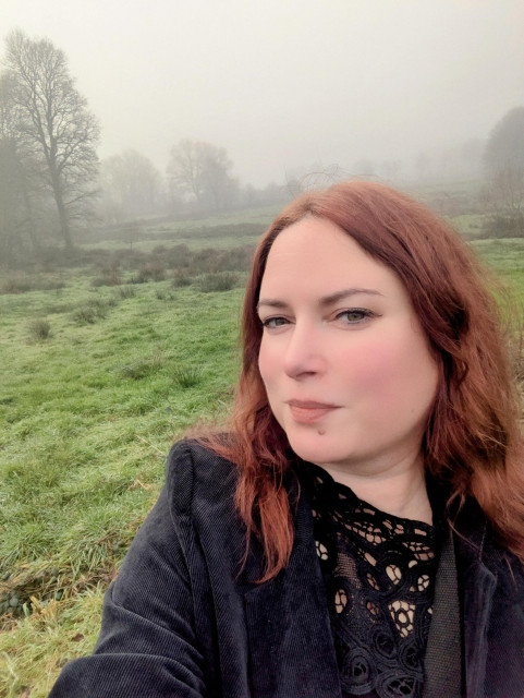 The image features a person with red-brown hair taking a selfie outdoors. They are wearing a black lace top under a dark jacket, and the background shows a misty, grassy field with scattered trees in a foggy setting, creating a serene and atmospheric scene.

