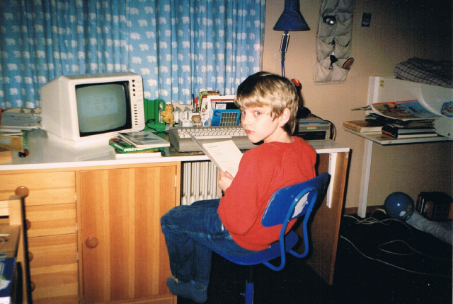 9-year old Jan sitting on his desk Xmas 1987. An ALDI C64 breadbin and a 1541 floppy disk drive are visible, connected to a cheap black and white television set. You can barely make out GEOS on the screen.