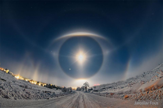 A snow covered road goes up a hill to a sky filled with stars. Arcs and halos in the sky ahead appear similar to a giant eye.