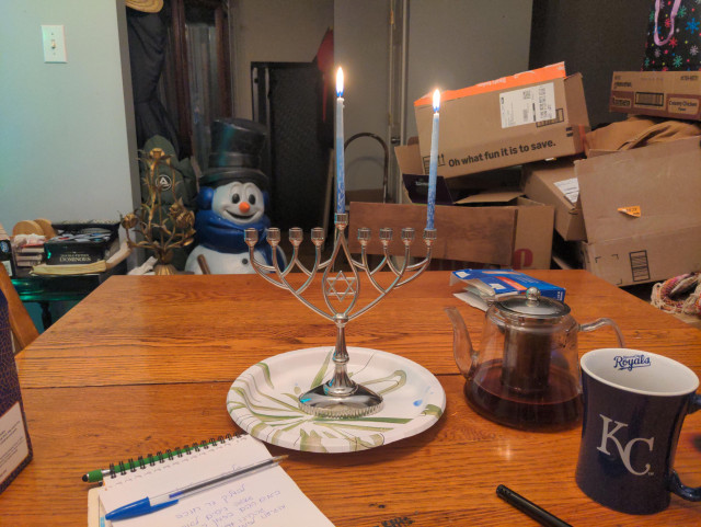 Wooden table with Hanukkah menorah with two candles. Notepad with handwritten Hebrew blessings. Kansas City Royals mug. Glass teapot. A bunch of stupid junk in the background.