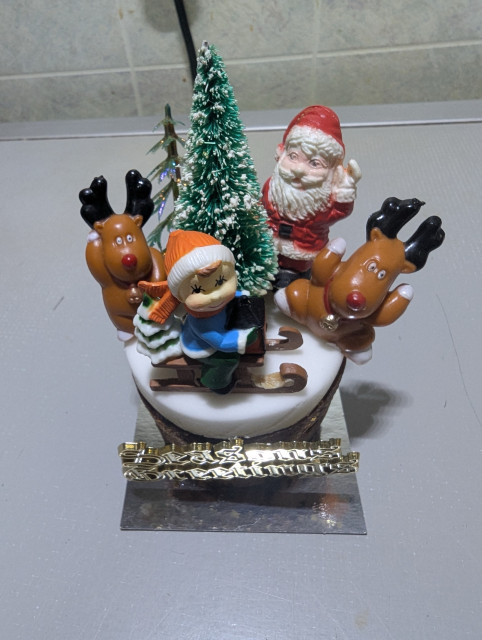 An individual Christmas cake decorated with a Santa Claus, a child on a sled, two reindeer, two small decorated pine trees, all set atop a white iced top with a gold inscription reading Season’s Greetings pinned to the side. 