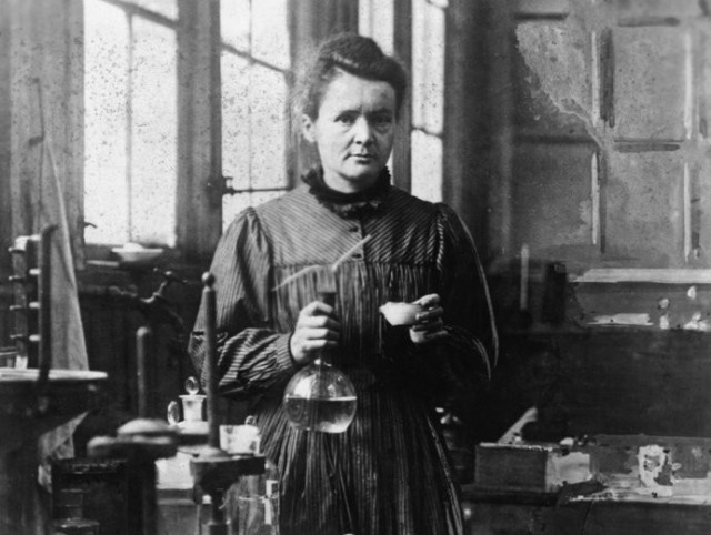 posed photograph of Marie Curie in her lab holding a volumetric flask. She is a white woman with dark hair.
