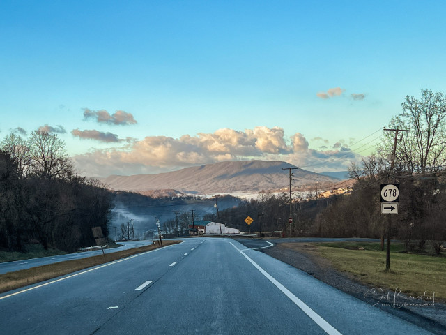 A scenic highway in Virginia under a bright blue sky, leading to distant mountains. Bare trees line the road, and wisps of mist rise from the valley, creating a peaceful, open-road vibe.